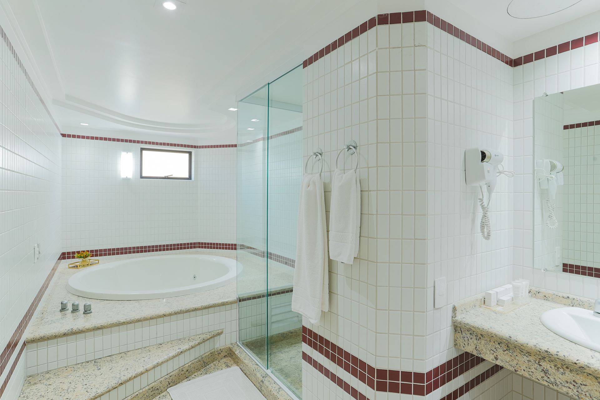 Banheiro branco e luminoso com banheira de canto, box de chuveiro revestido com azulejos e detalhes em vermelho.