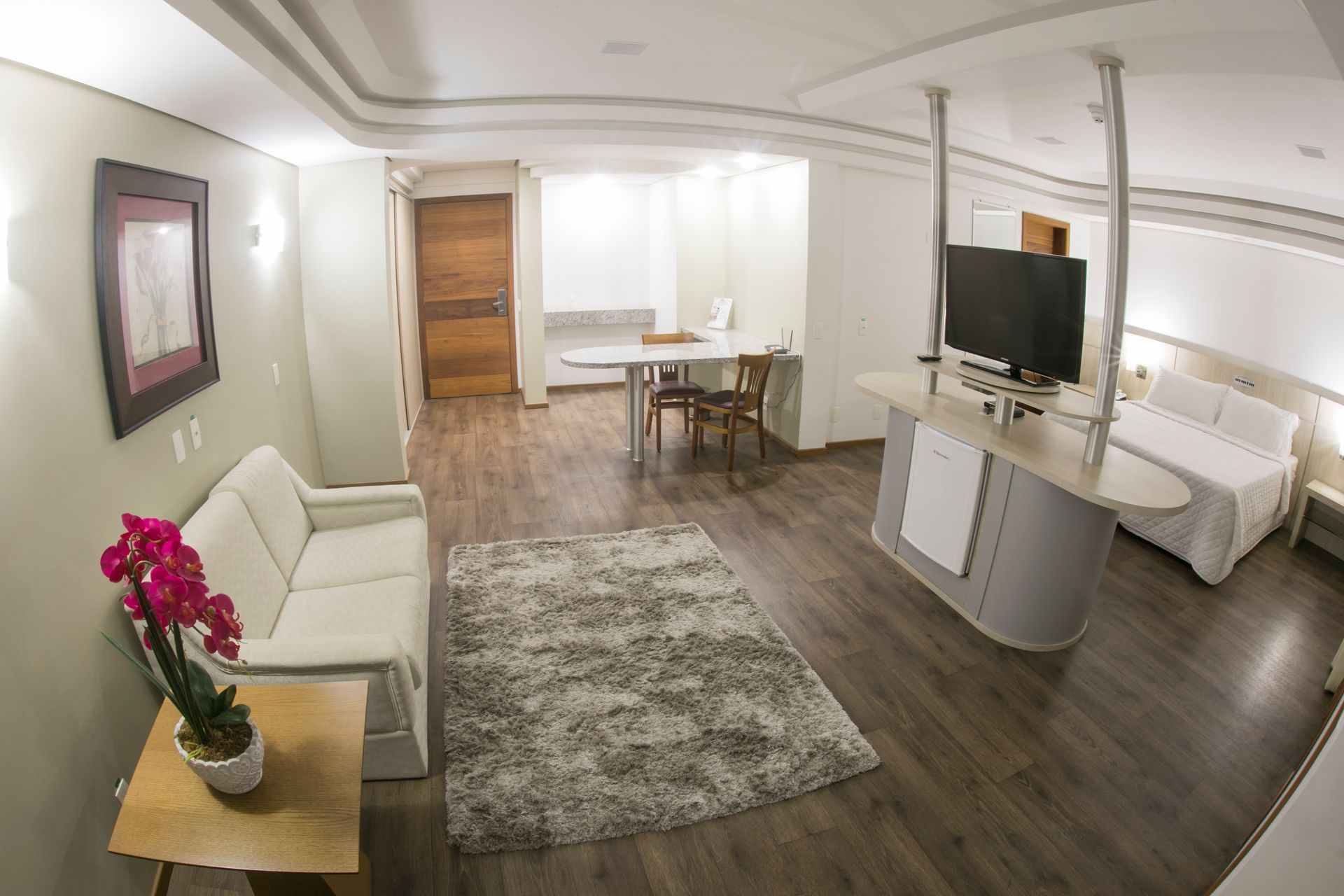 Apartamento estúdio moderno com sofá, cama, escrivaninha, TV e área para refeições em piso de madeira.