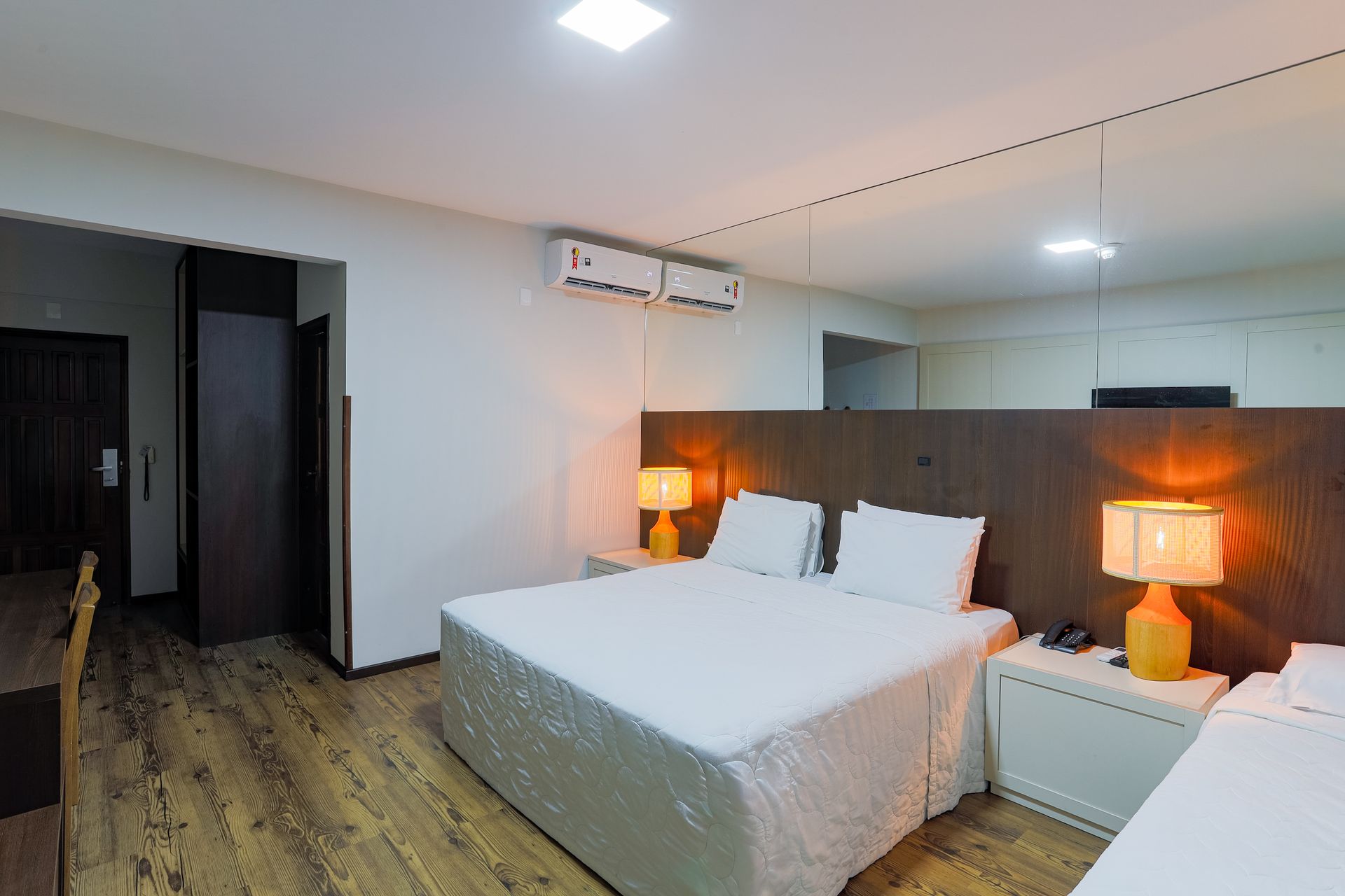 Quarto de hotel moderno com duas camas, piso de madeira, parede espelhada e abajures aconchegantes.