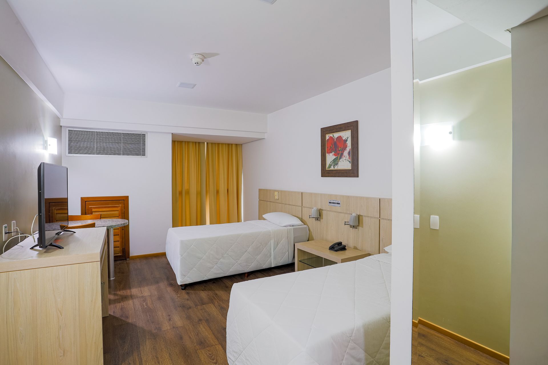 Quarto de hotel moderno com duas camas, piso de madeira, cortinas amarelas e uma pequena cozinha.