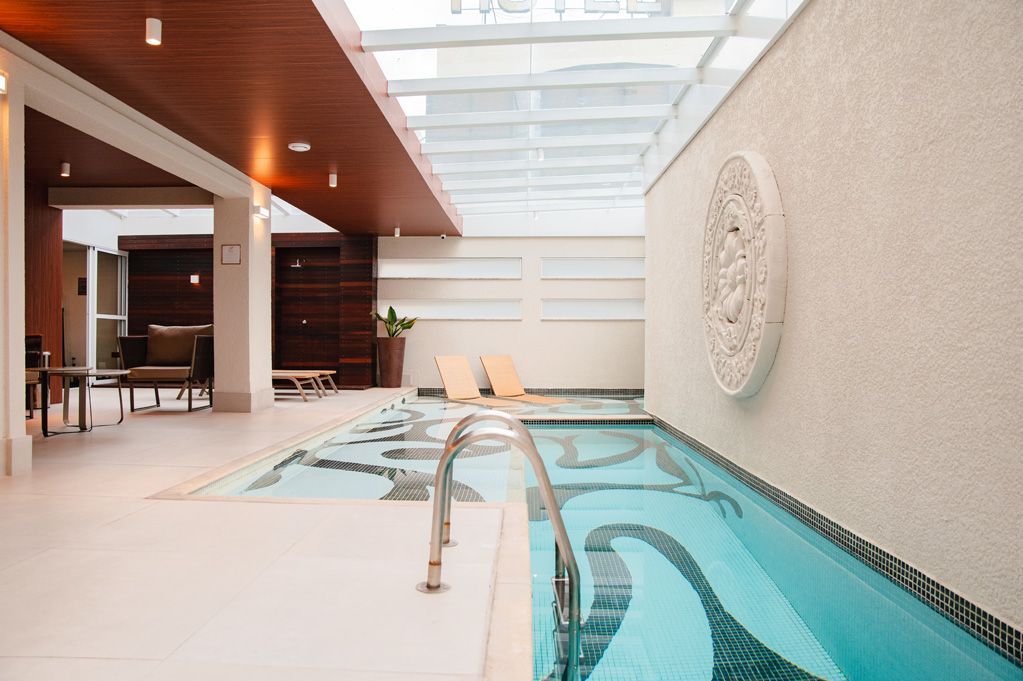 Piscina interior com água cristalina, teto com claraboia e área de estar estilo spa ao lado.