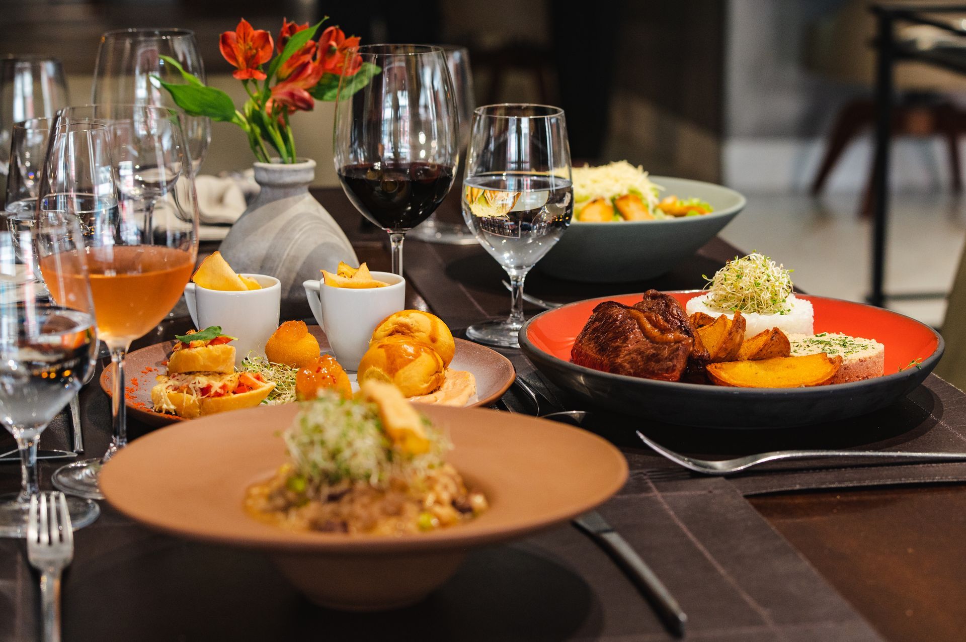 Mesa de jantar elegante com vinho, coquetéis e pratos gourmet em um ambiente de restaurante.