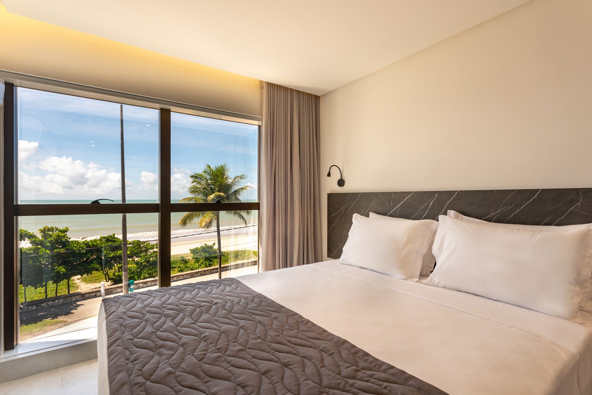 Quarto de hotel moderno com janela ampla, vista para o mar e cama arrumada.
