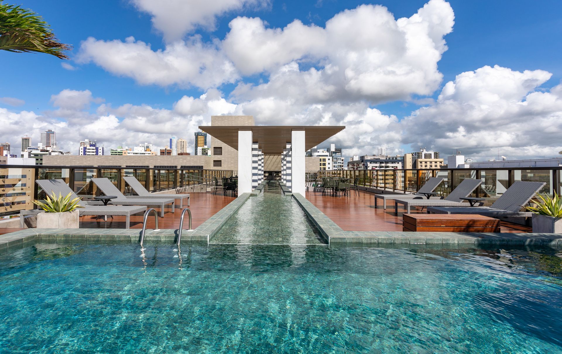 Piscina na cobertura com água turquesa, espreguiçadeiras e vista para o horizonte da cidade sob um céu azul nublado.