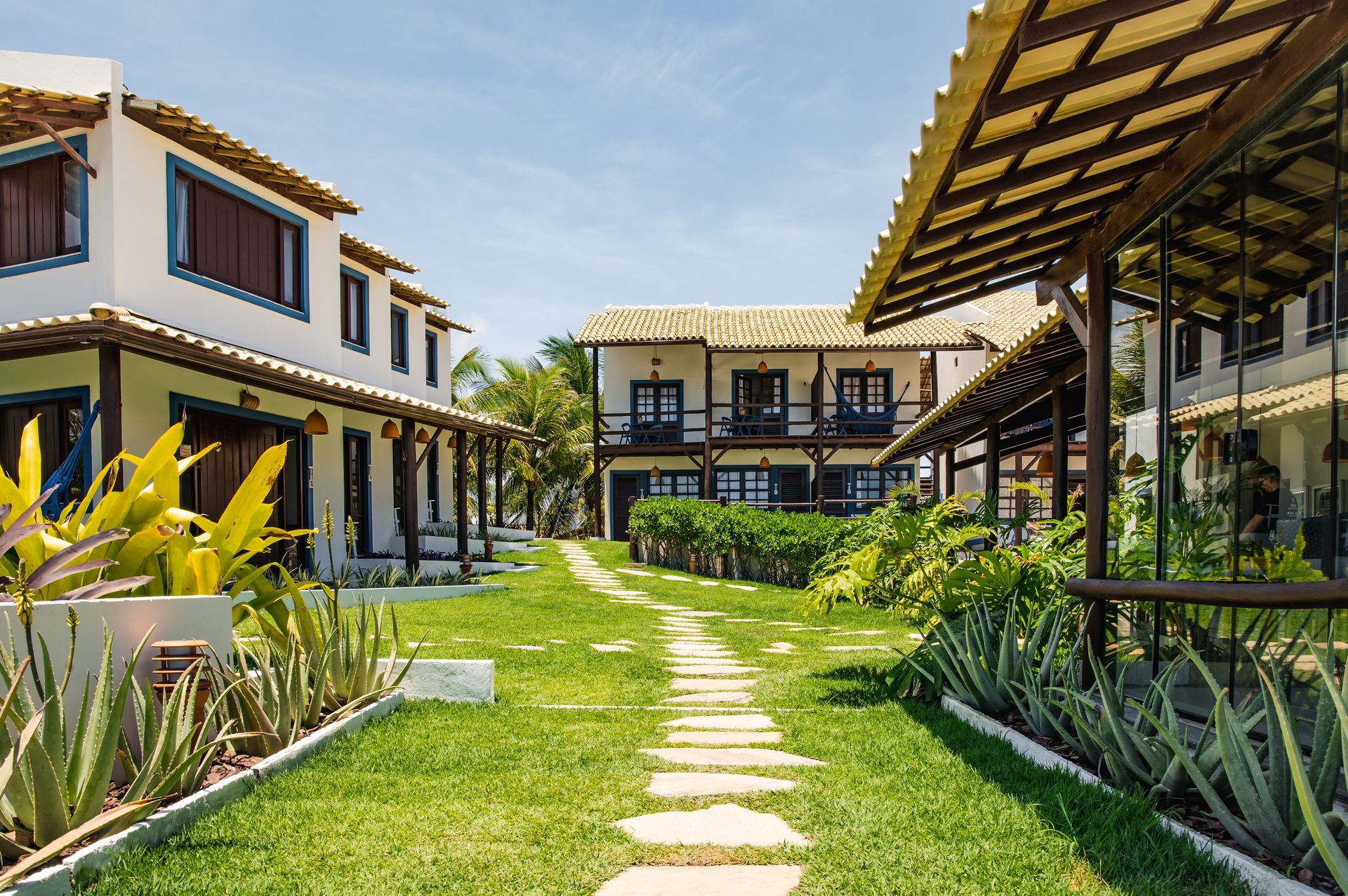 Pátio de resort moderno com gramado verde, caminho de pedra e edifícios brancos sob um céu azul brilhante.