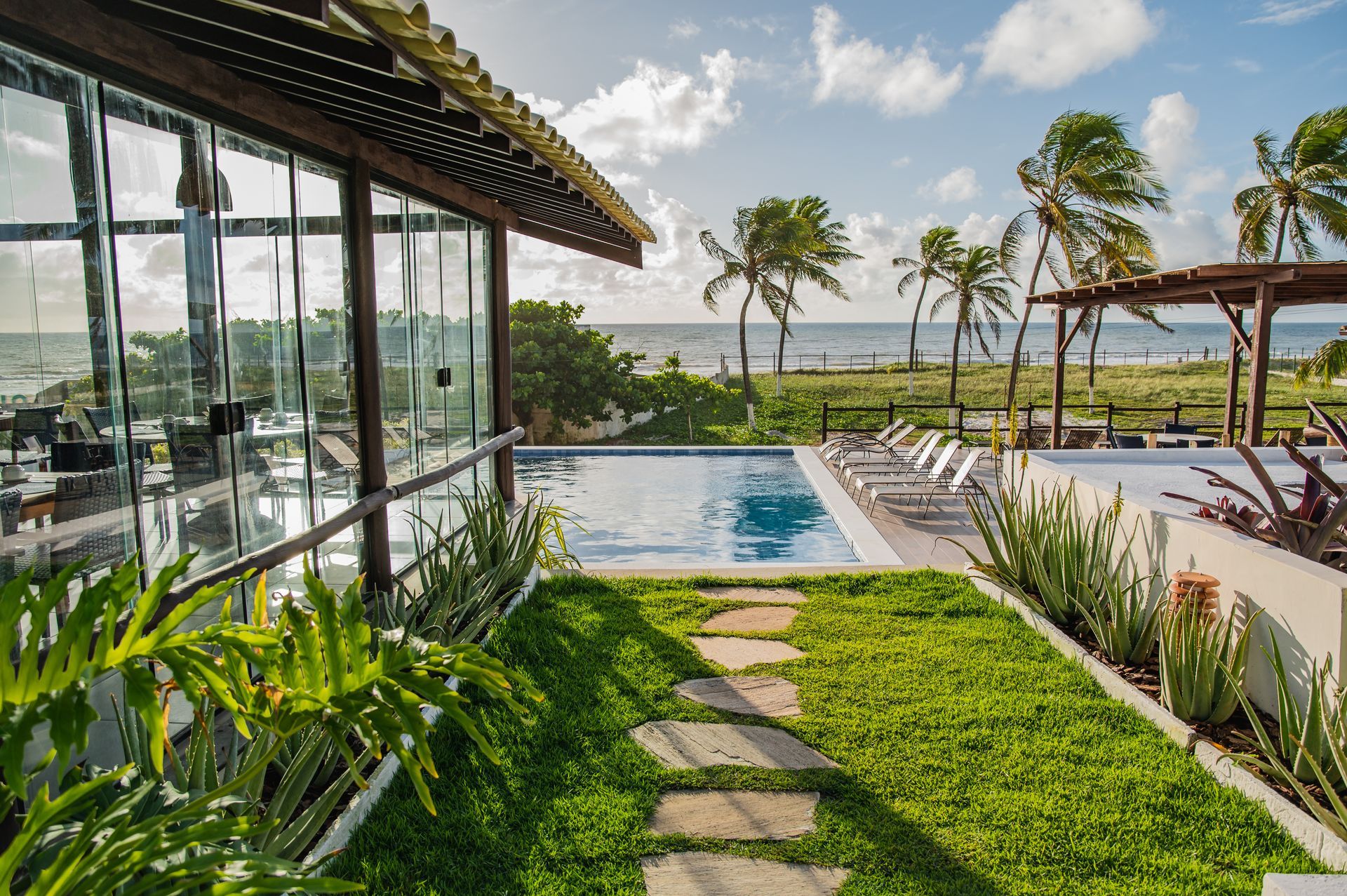 Quintal moderno com piscina, pátio envidraçado, gramado exuberante, palmeiras e vista para o mar.