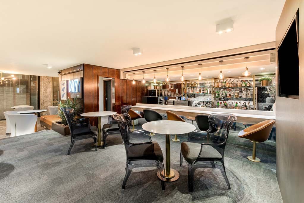 Lounge bar moderno com mesas, cadeiras pretas, balcão de mármore e decoração em madeira e vidro em tons quentes.