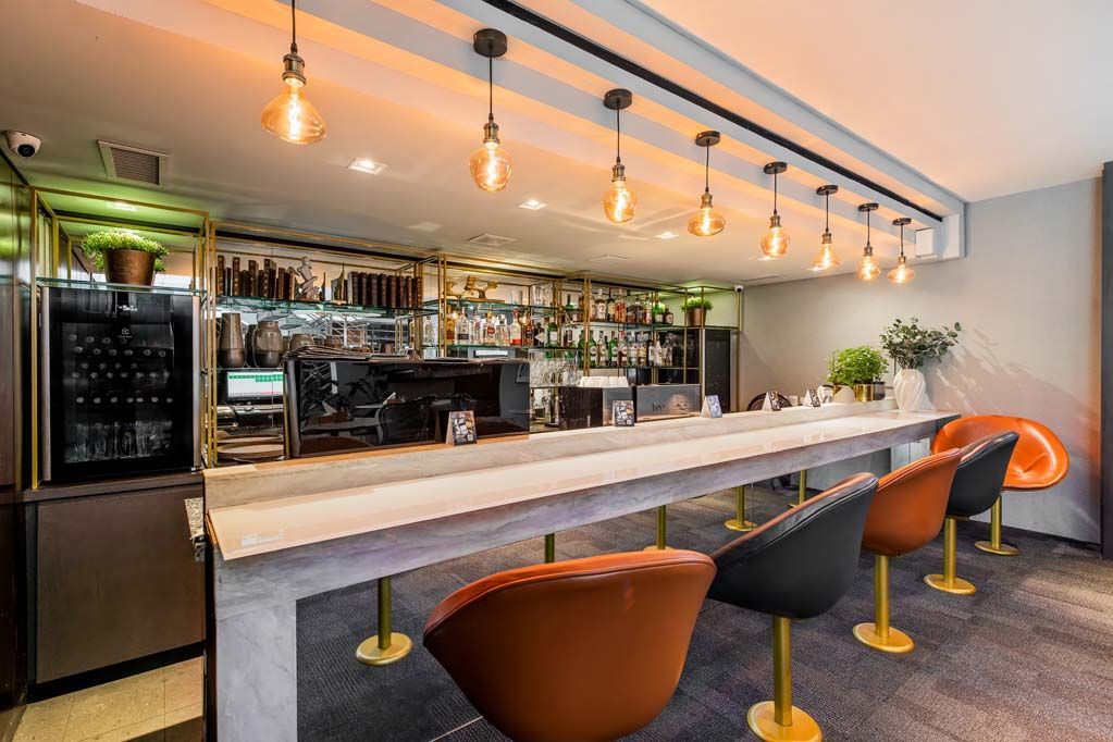 Interior de bar moderno com um longo balcão branco, luminárias pendentes e banquetas marrons e laranjas.