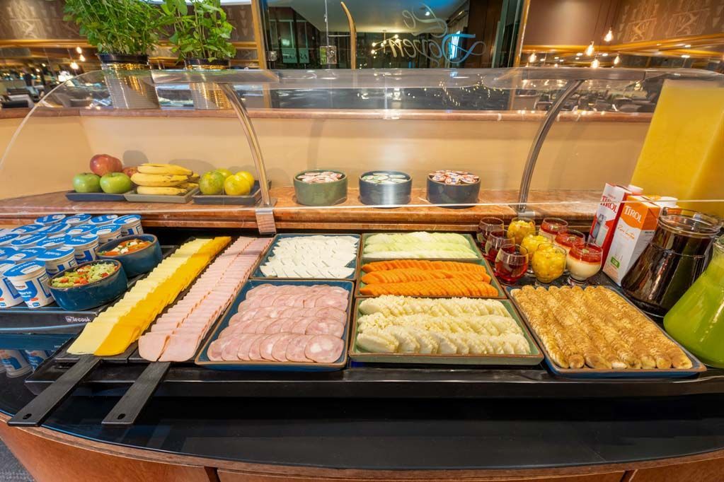 Balcão de buffet com bandejas de alimentos variados, incluindo legumes, carnes e molhos, sob uma proteção de vidro contra espirros.