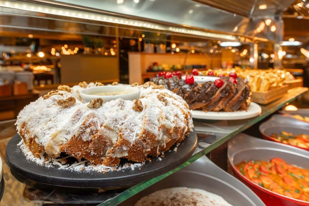 Doces expostos em uma vitrine de padaria, incluindo um bolo Bundt polvilhado com açúcar e tortas, sob luzes aconchegantes.