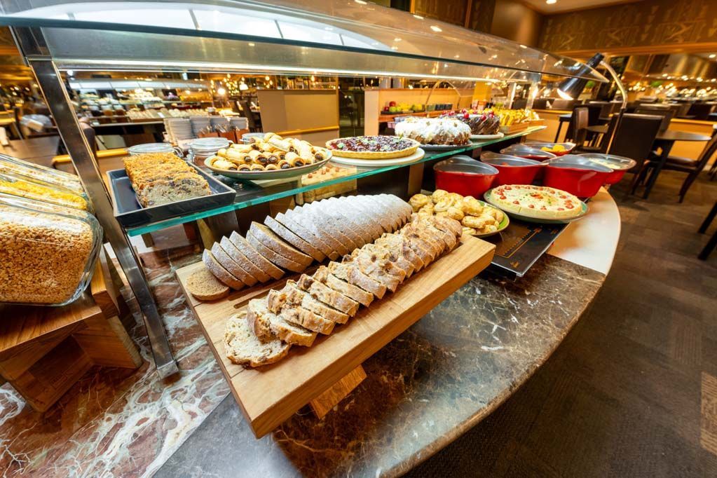 Buffet de hotel com bandejas de doces, pães, frutas e pratos de café da manhã em um balcão polido.