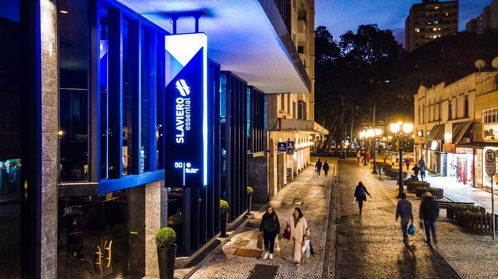 Cena noturna de rua com pessoas caminhando por uma calçada de paralelepípedos ao lado de prédios iluminados e uma placa azul brilhante.