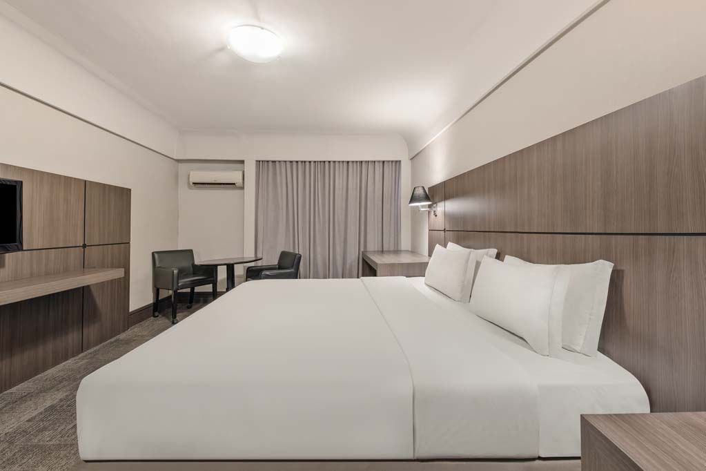 Quarto de hotel moderno com cama grande, console de TV, cadeiras de escritório e janela com cortina.