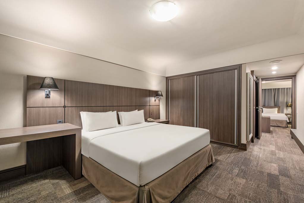 Quarto de hotel moderno com cama king-size, penteadeira, armário com espelho e corredor acarpetado que leva ao banheiro.