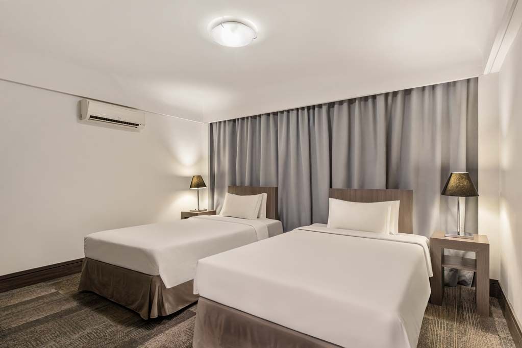 Quarto de hotel com duas camas de solteiro, cortinas cinza, abajures e piso acarpetado.