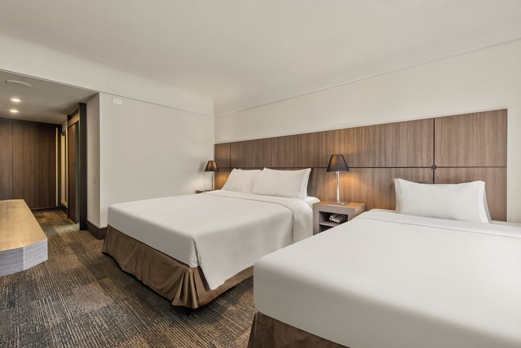 Quarto de hotel moderno com duas camas brancas, cabeceira de madeira e abajures aconchegantes.