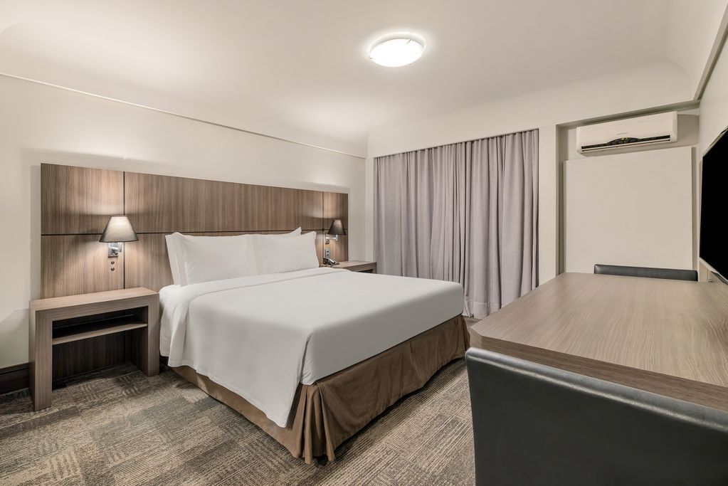 Quarto de hotel impecavelmente arrumado com cama branca, cortinas cinza, abajur e escrivaninha.
