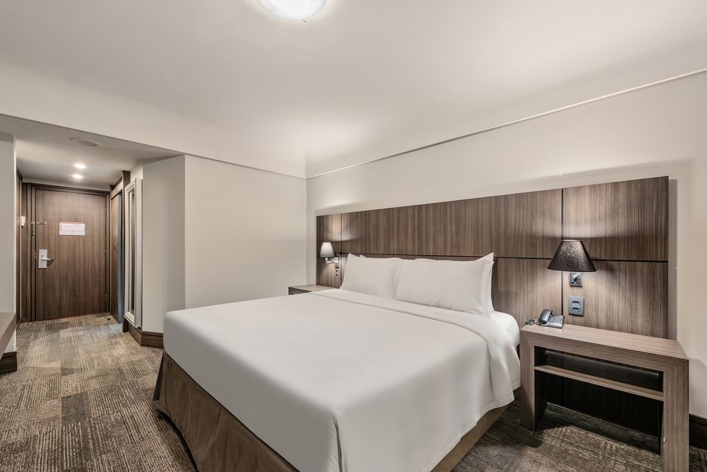 Quarto de hotel moderno com cama grande, cabeceira de madeira, mesa de cabeceira e entrada pelo corredor.