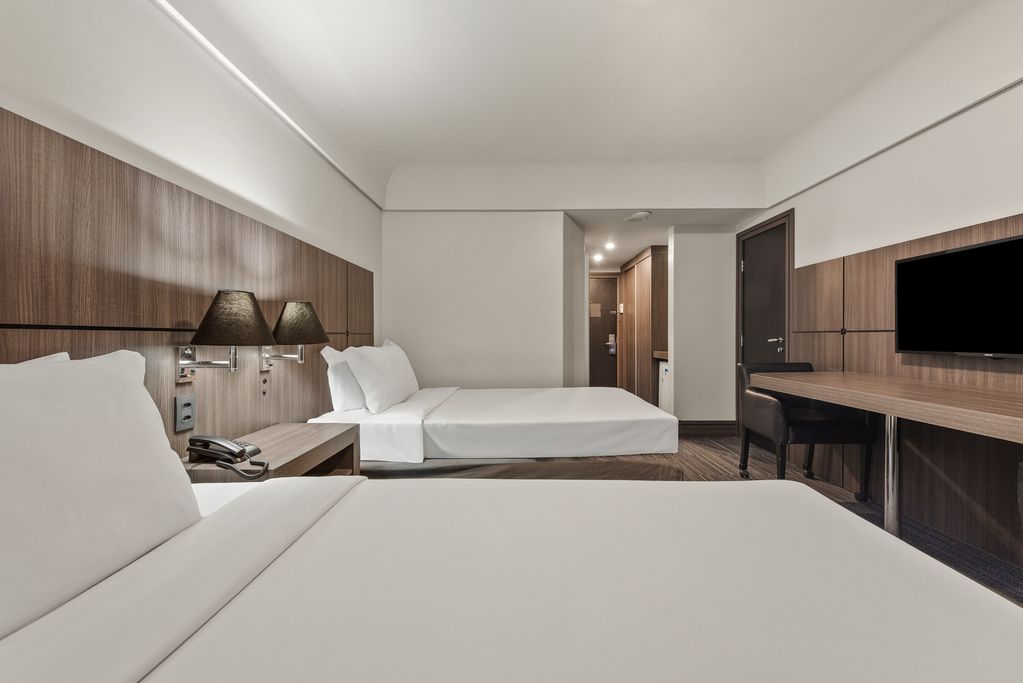 Quarto de hotel moderno com duas camas, parede revestida de madeira, escrivaninha e TV de parede.