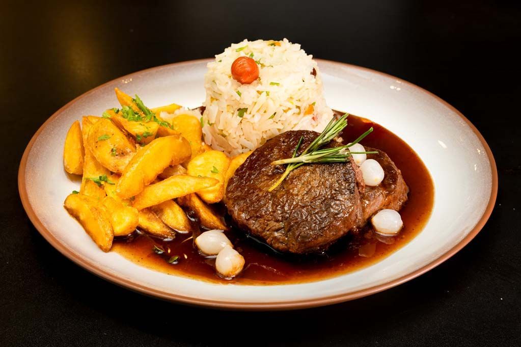 Bife com molho, arroz e batatas fritas em um prato branco.
