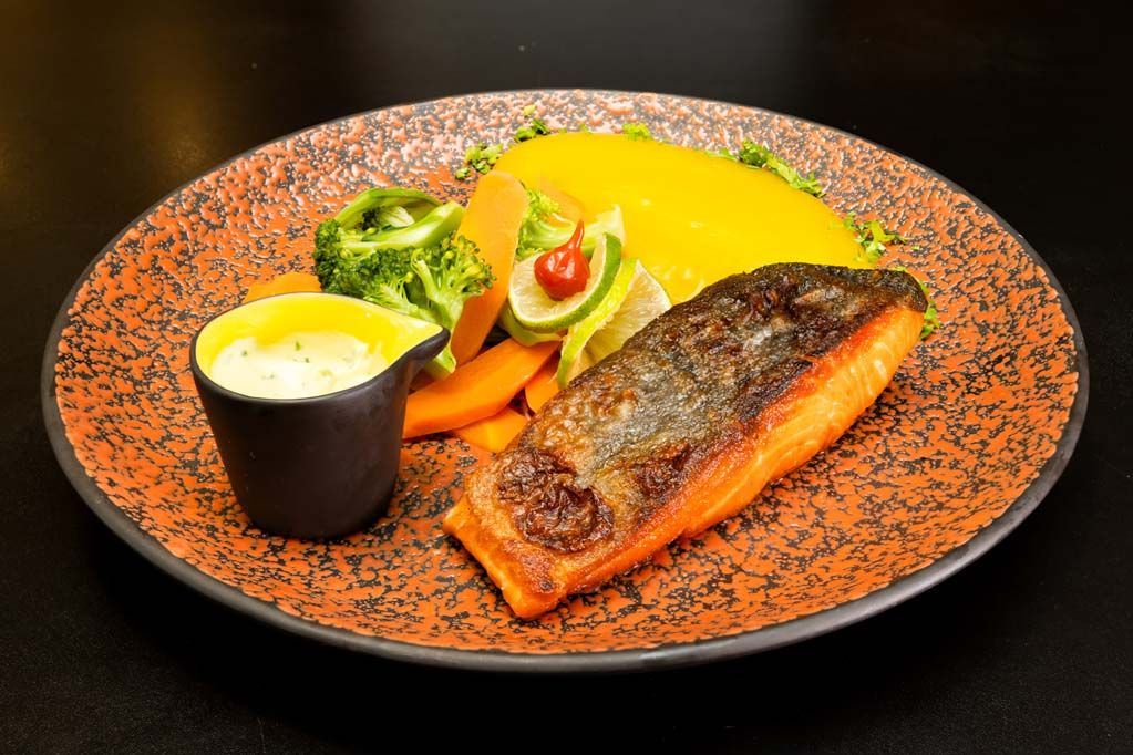 Salmão grelhado com legumes, molho amarelo e purê de batatas em um prato com pintas laranja.