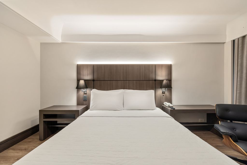 Quarto de hotel moderno com cama arrumada, mesas de cabeceira e luminária de parede.
