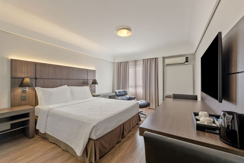 Quarto de hotel moderno com cama grande, escrivaninha, TV de parede e janela com cortina.