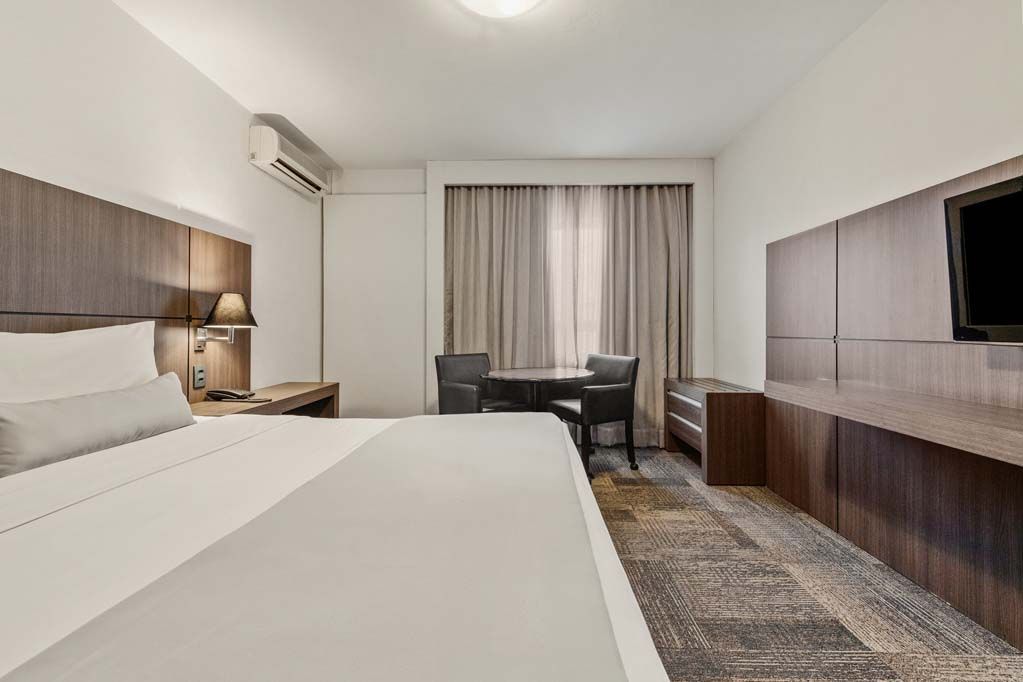Quarto de hotel moderno com cama, escrivaninha, TV de parede e cortinas junto à janela.