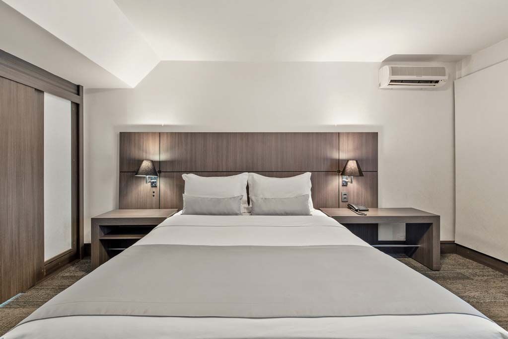 Quarto de hotel moderno com cama grande, cabeceira de madeira, abajures e ar-condicionado de parede.