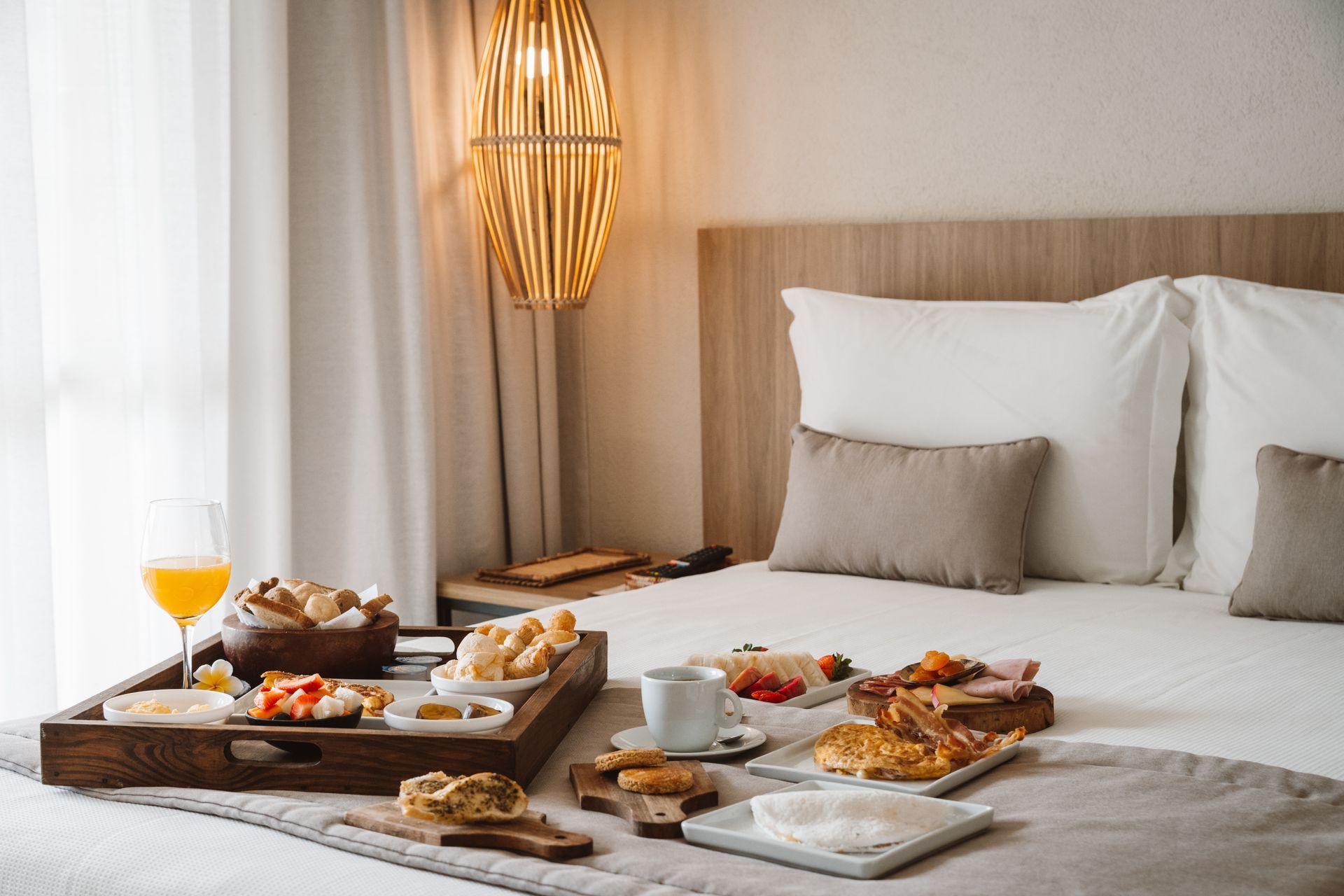 Bandeja de café da manhã sobre a cama em um quarto de hotel iluminado, com café, suco de laranja, doces e pratos.