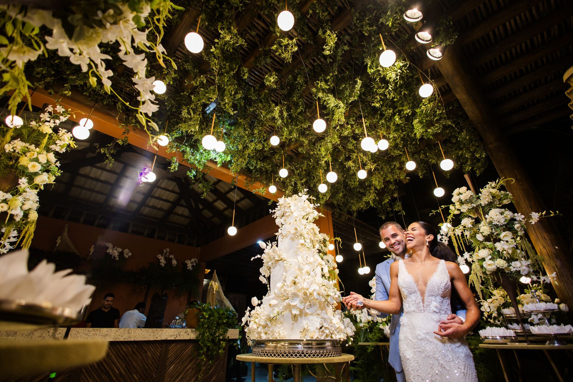 Duas pessoas de vestido branco em uma recepção de casamento iluminada por flores, ao lado de um bolo alto sob luzes pendentes.