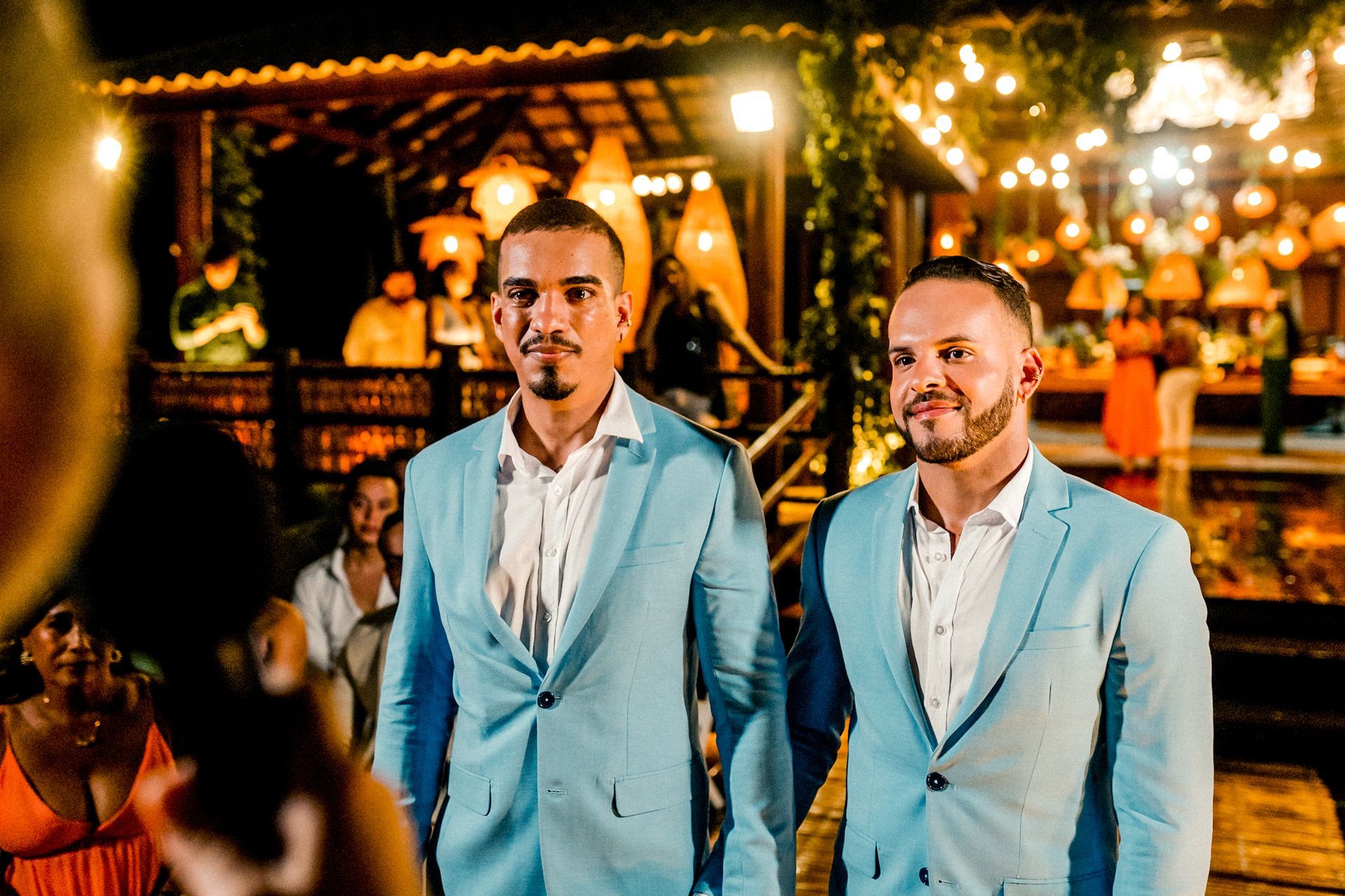 Dois homens de terno azul-claro em pé ao ar livre em um evento noturno com iluminação aconchegante.