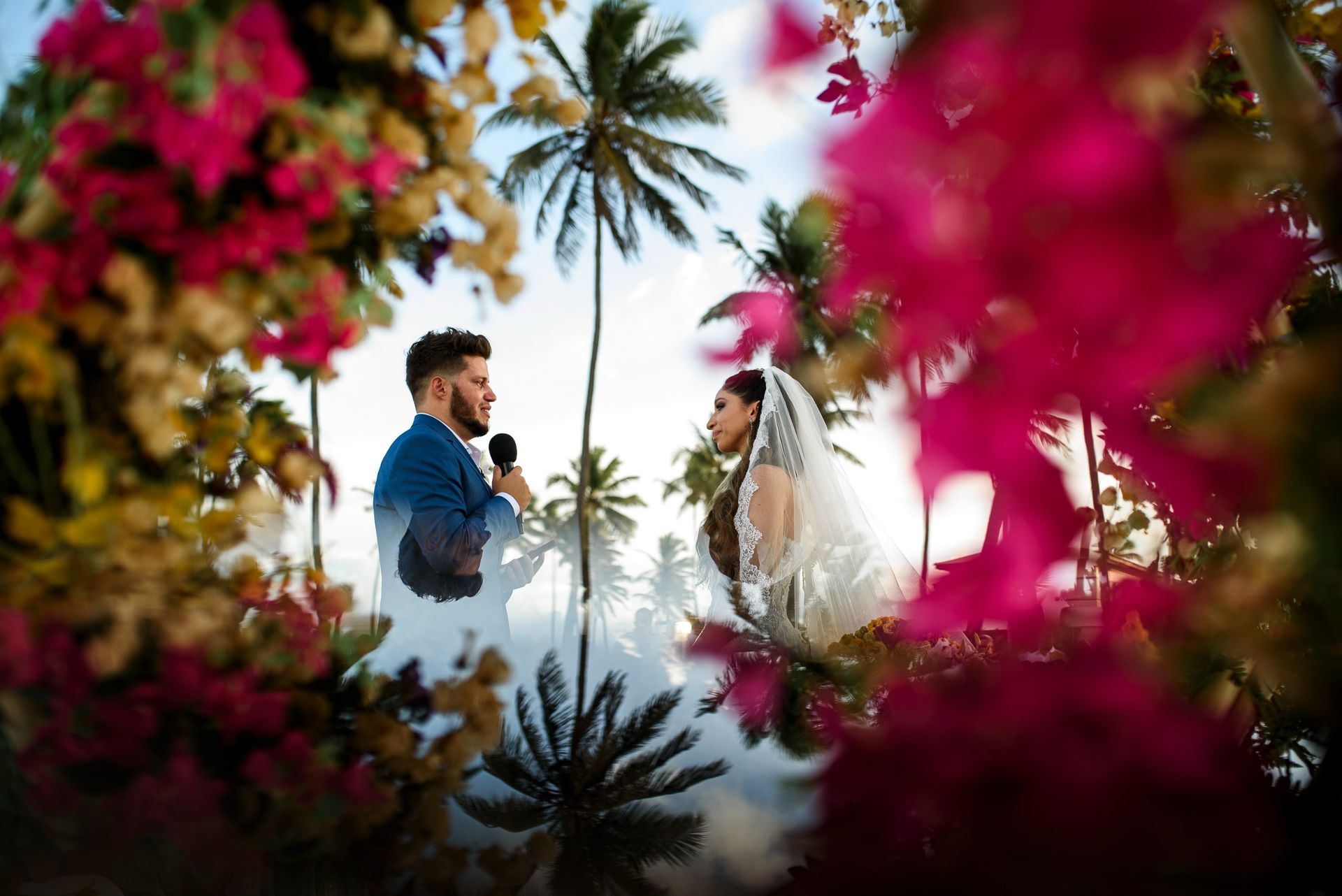 Casal de noivos frente a frente ao ar livre, emoldurados por flores cor-de-rosa e palmeiras.