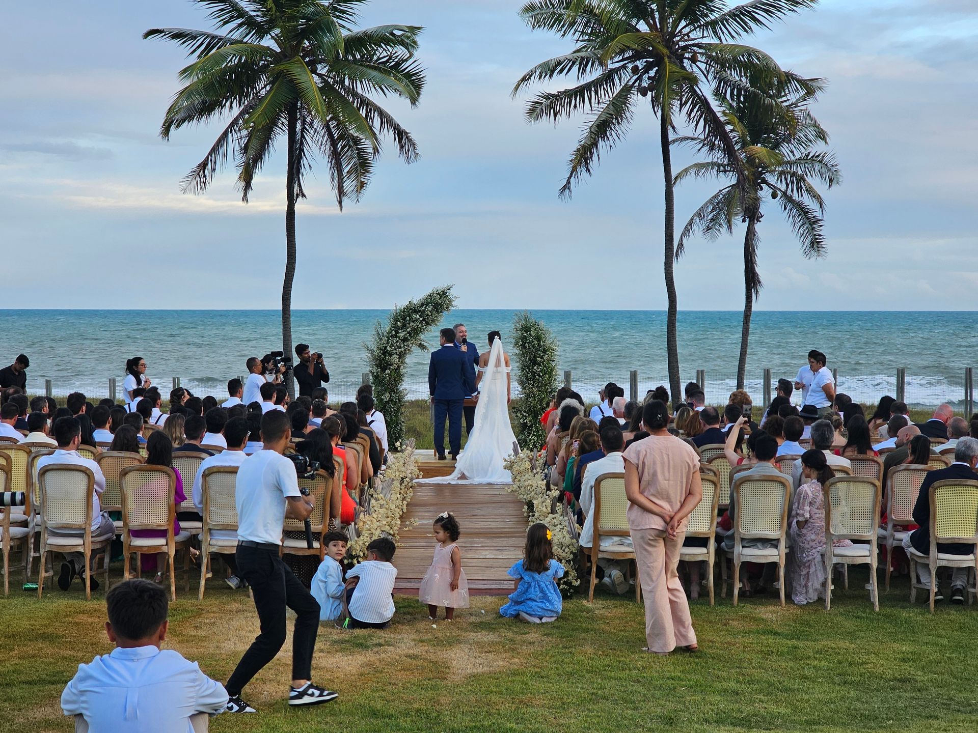 Cerimônia de casamento ao ar livre na praia, com os convidados sentados na grama de frente para o corredor que leva ao oceano.