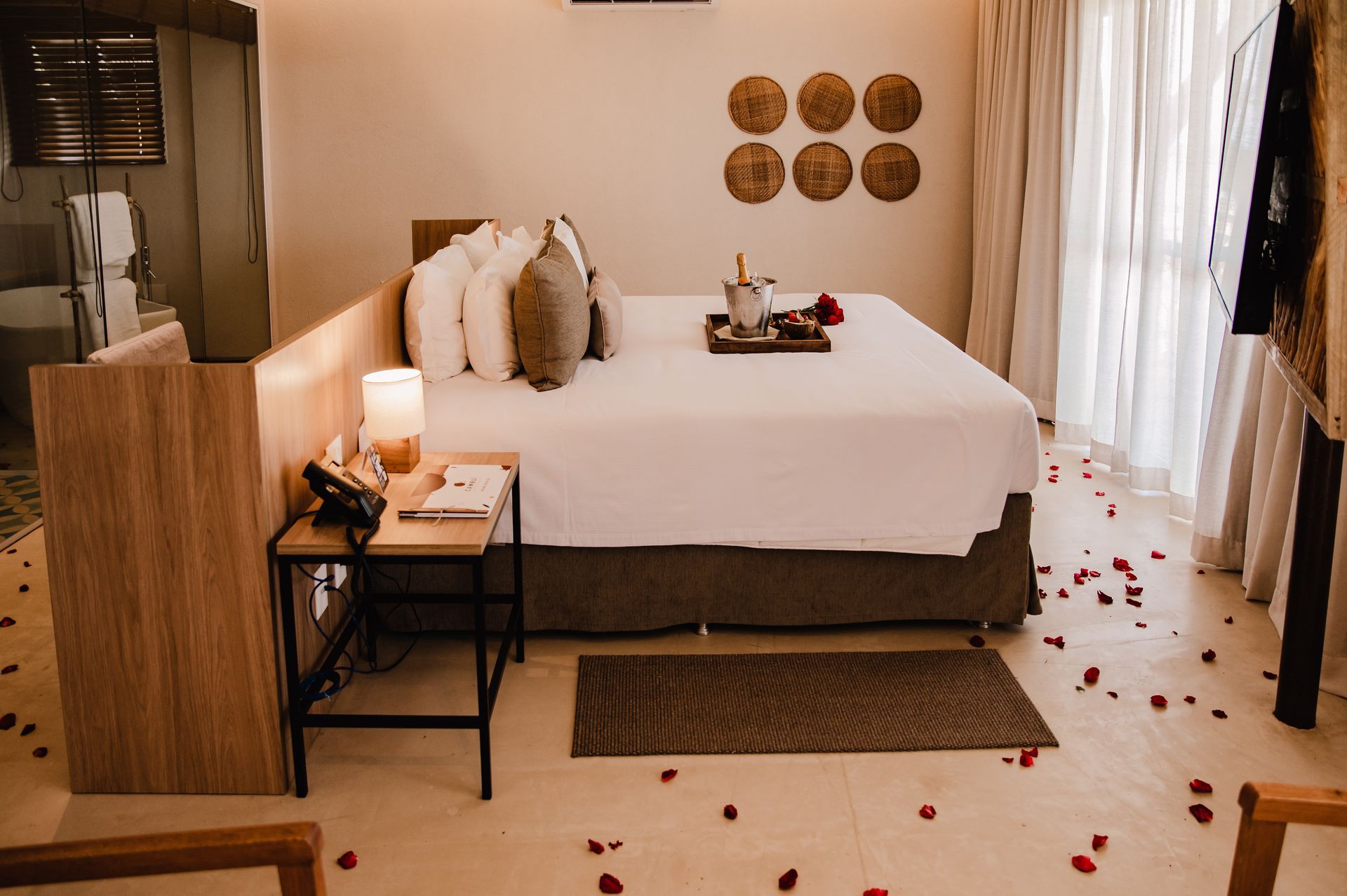 Quarto aconchegante com cama arrumada, mesa de cabeceira, pétalas de rosa no chão e iluminação suave e quente.
