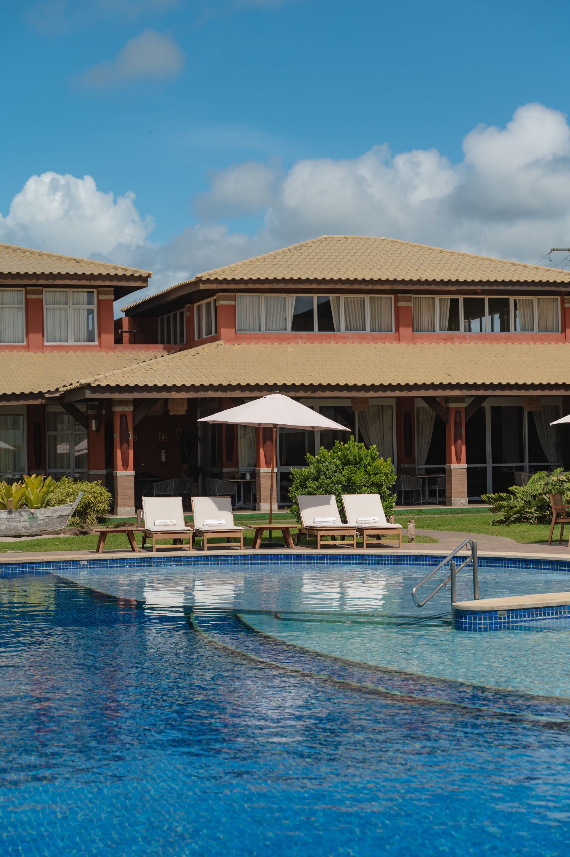 Piscina de resort de luxo com espreguiçadeiras e um prédio de dois andares sob um céu azul.