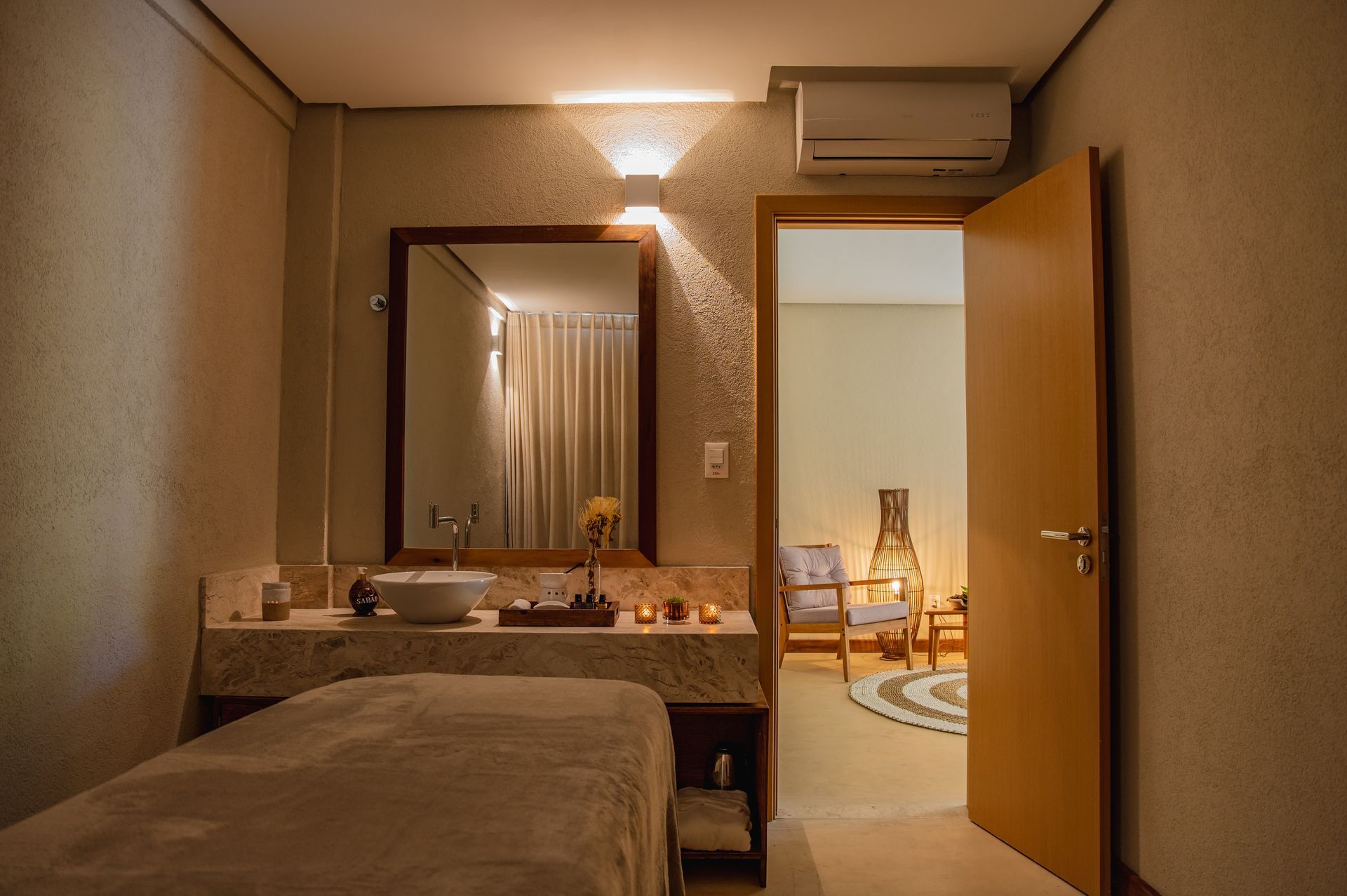 Quarto de hotel aconchegante com cama, espelho de maquilhagem e porta aberta para uma área de estar iluminada.