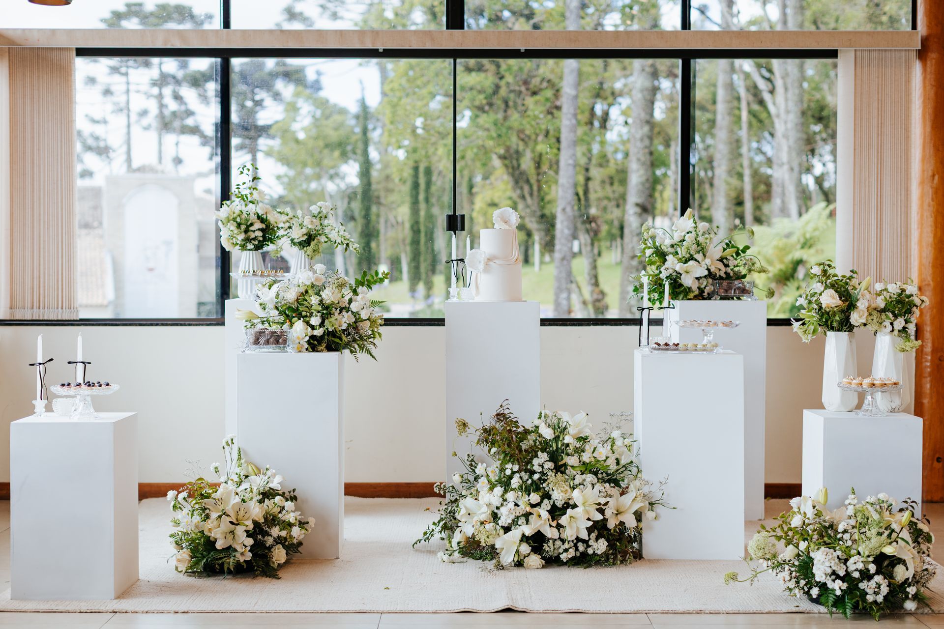 Arranjo floral branco para casamento, com flores em pedestais em frente a grandes janelas.