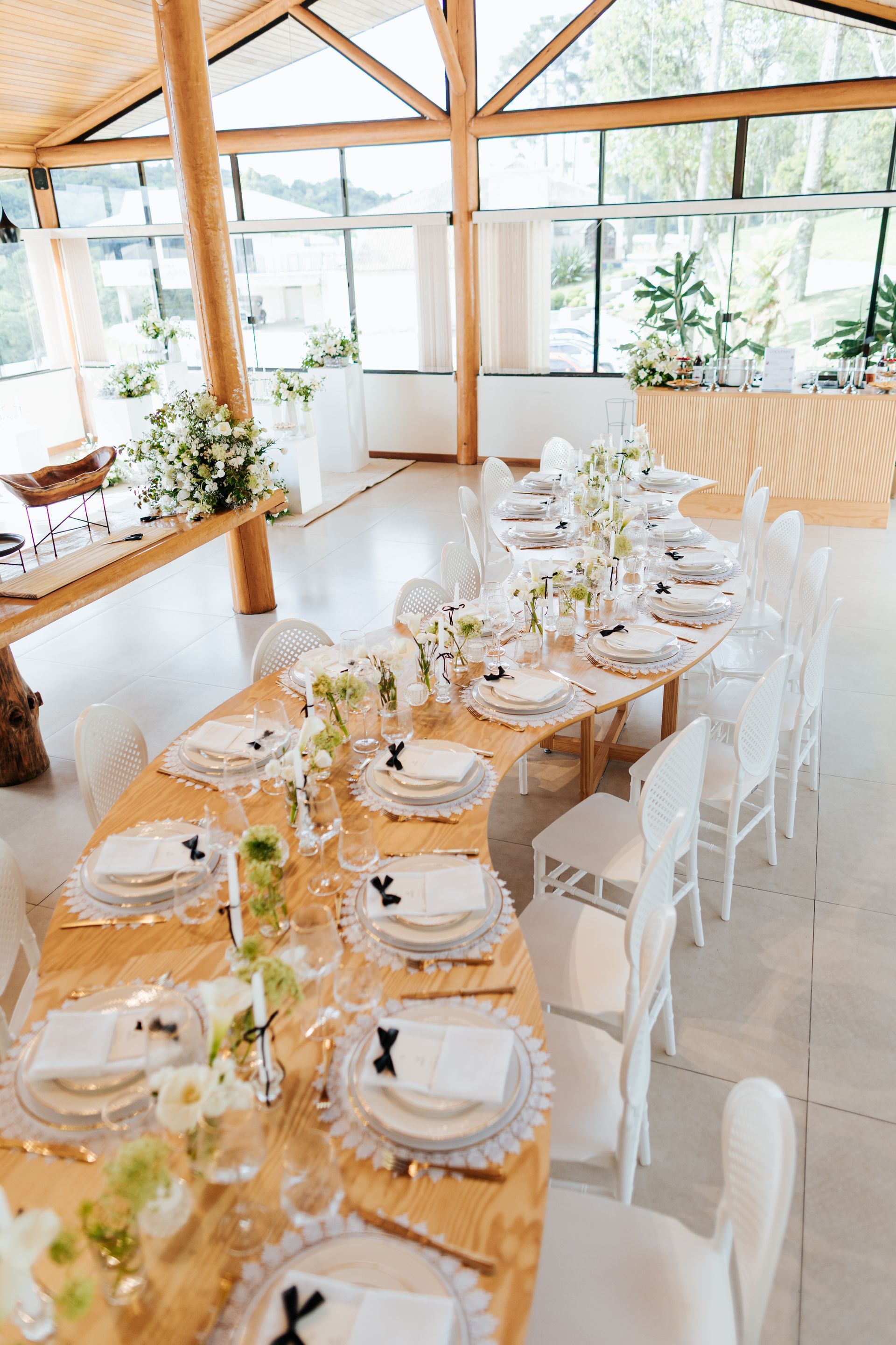 Mesa de jantar bem iluminada com cadeiras brancas em uma sala com paredes de vidro, decorada com plantas e utensílios de mesa.