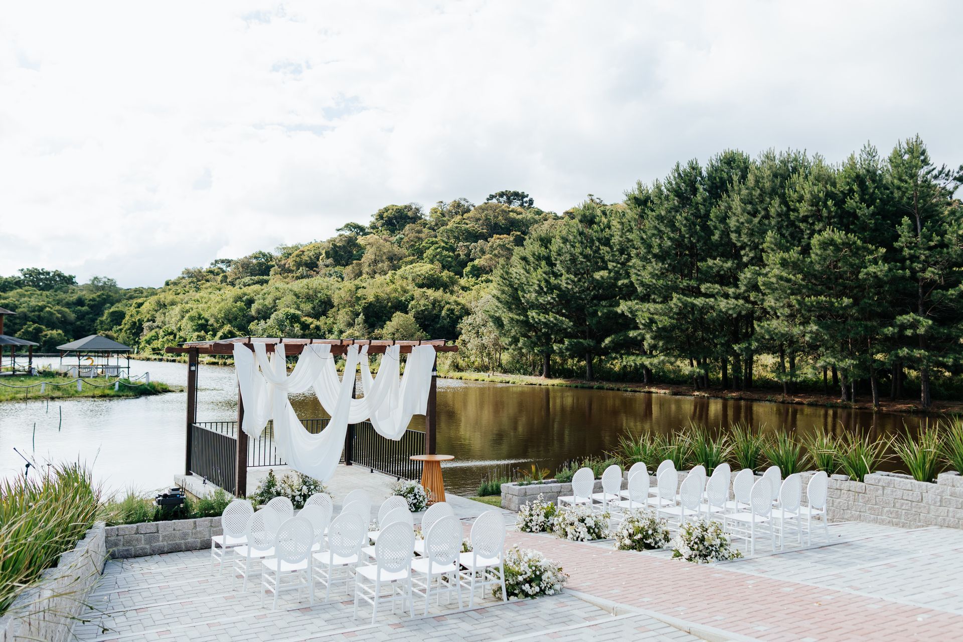 Decoração de casamento ao ar livre à beira de um lago com cadeiras brancas, arco drapeado e arranjos florais ao lado de árvores.