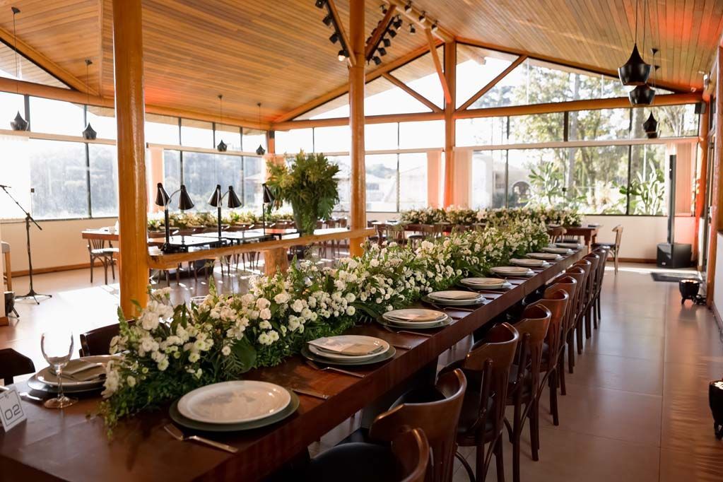 Elegante mesa de jantar comprida em uma sala iluminada com vigas de madeira, grandes janelas e arranjos de flores brancas como centro de mesa.