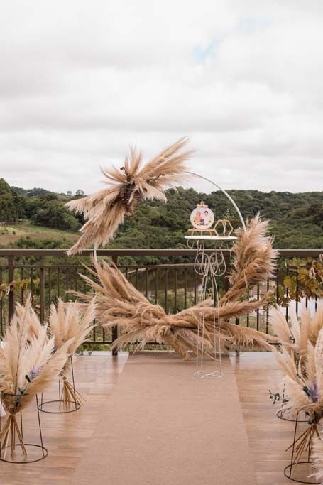Corredor de casamento ao ar livre decorado com capim-dos-pampas bege, arco e colinas pitorescas ao fundo.