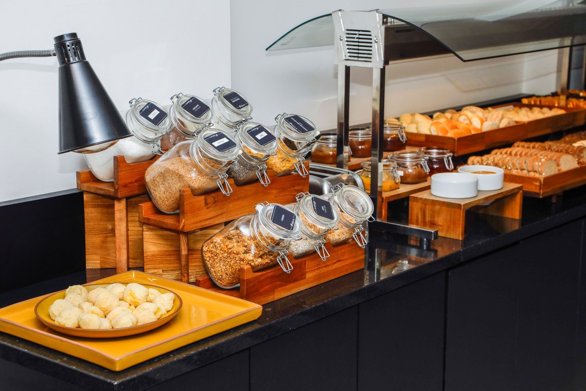 Buffet de padaria com cestas de pão, doces embalados e um prato de pãezinhos sob aquecedores.