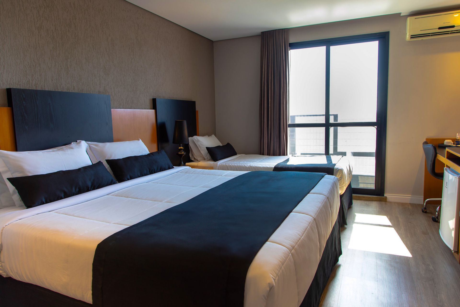 Quarto de hotel moderno com duas camas de solteiro, peseiras escuras e porta para varanda com vista para a cidade.