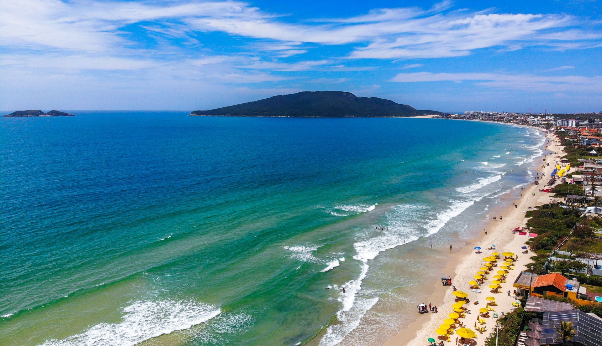 Vista aérea de uma praia azul-esverdeada com ondas, guarda-sóis amarelos e uma ilha distante no horizonte.