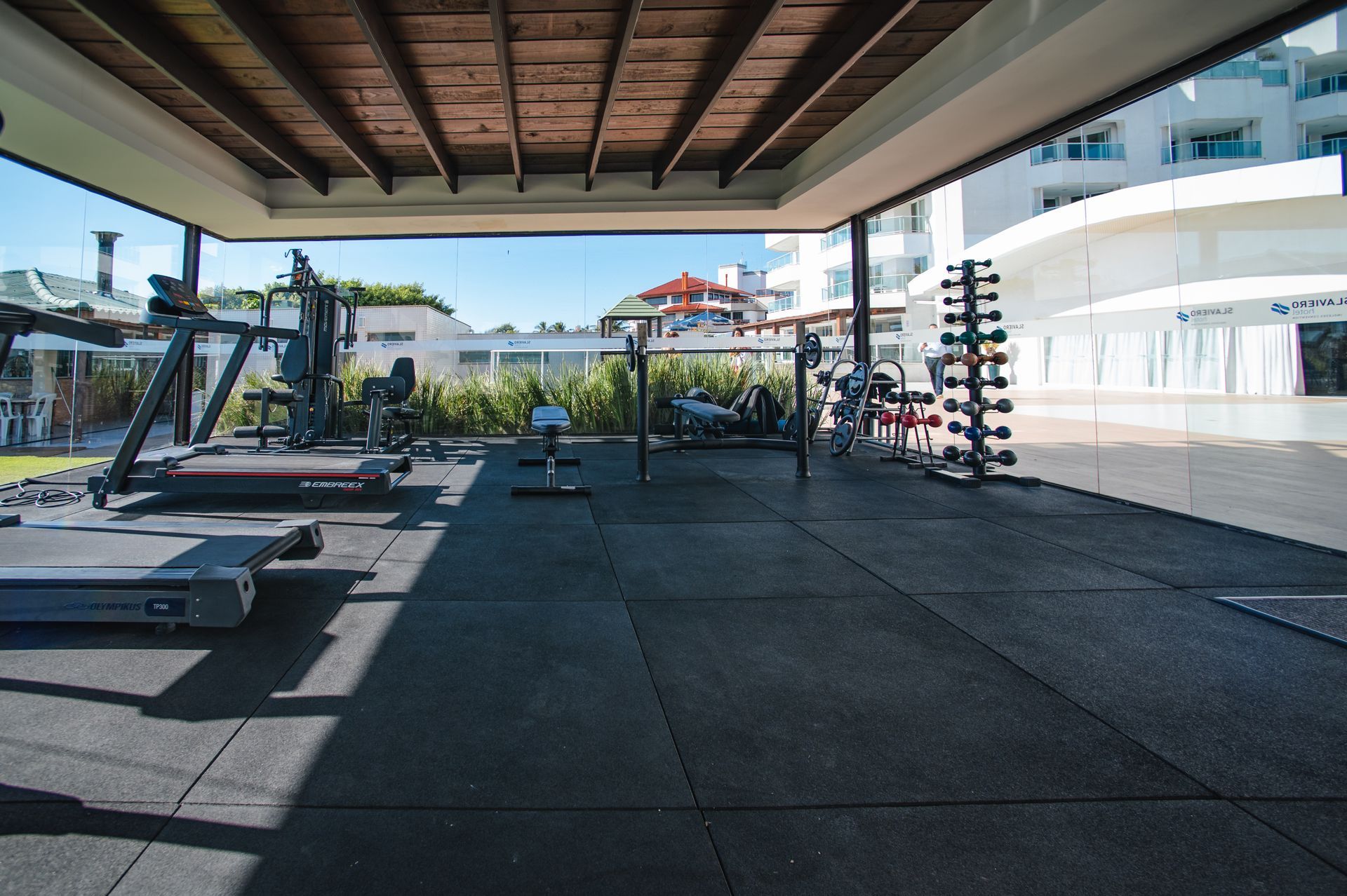 Academia ao ar livre com equipamentos de ginástica sob um pátio coberto, com vista para edifícios brancos modernos.