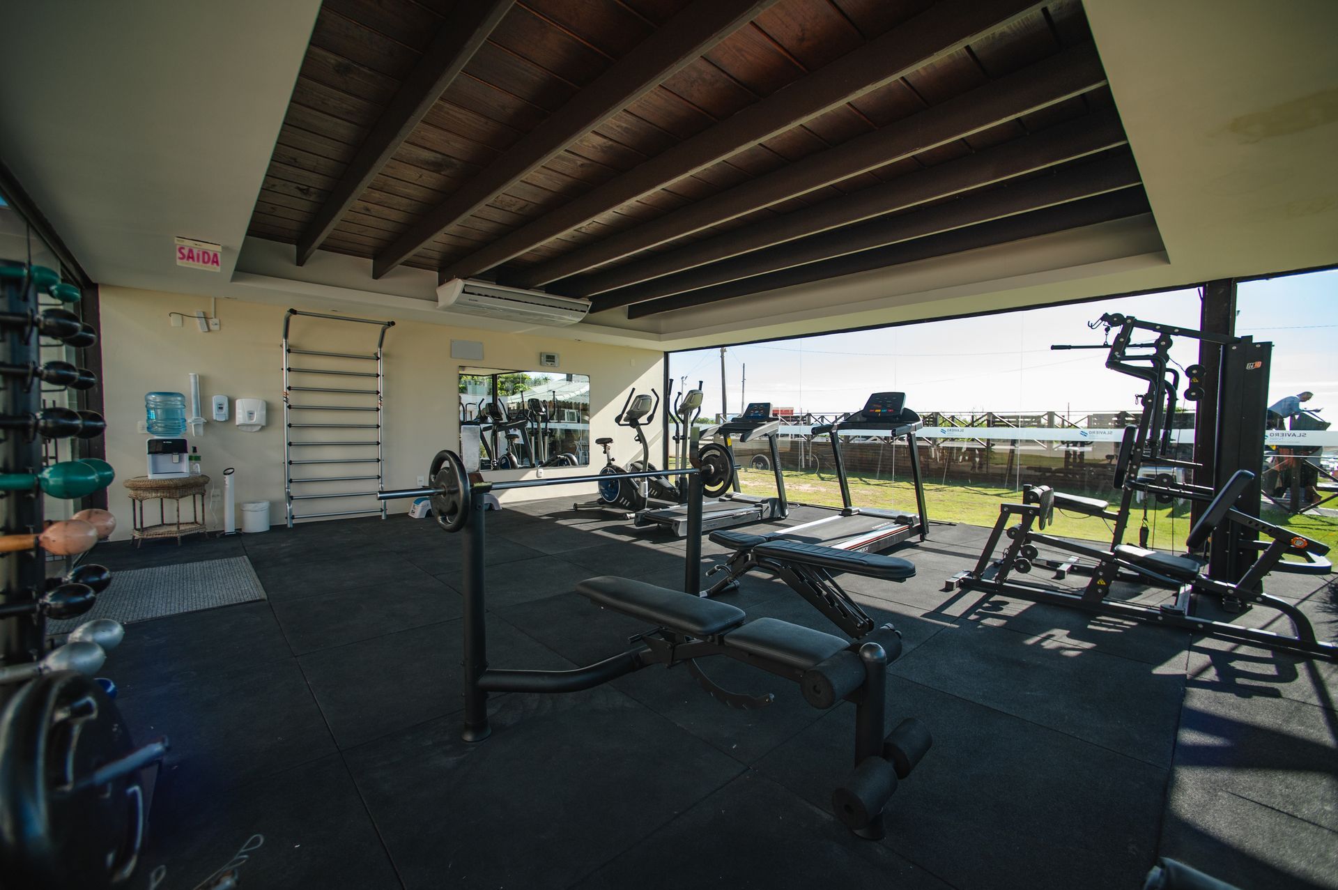 Academia ao ar livre sob um pátio coberto, com bancos de musculação, suportes e equipamentos de ginástica, à luz do dia.