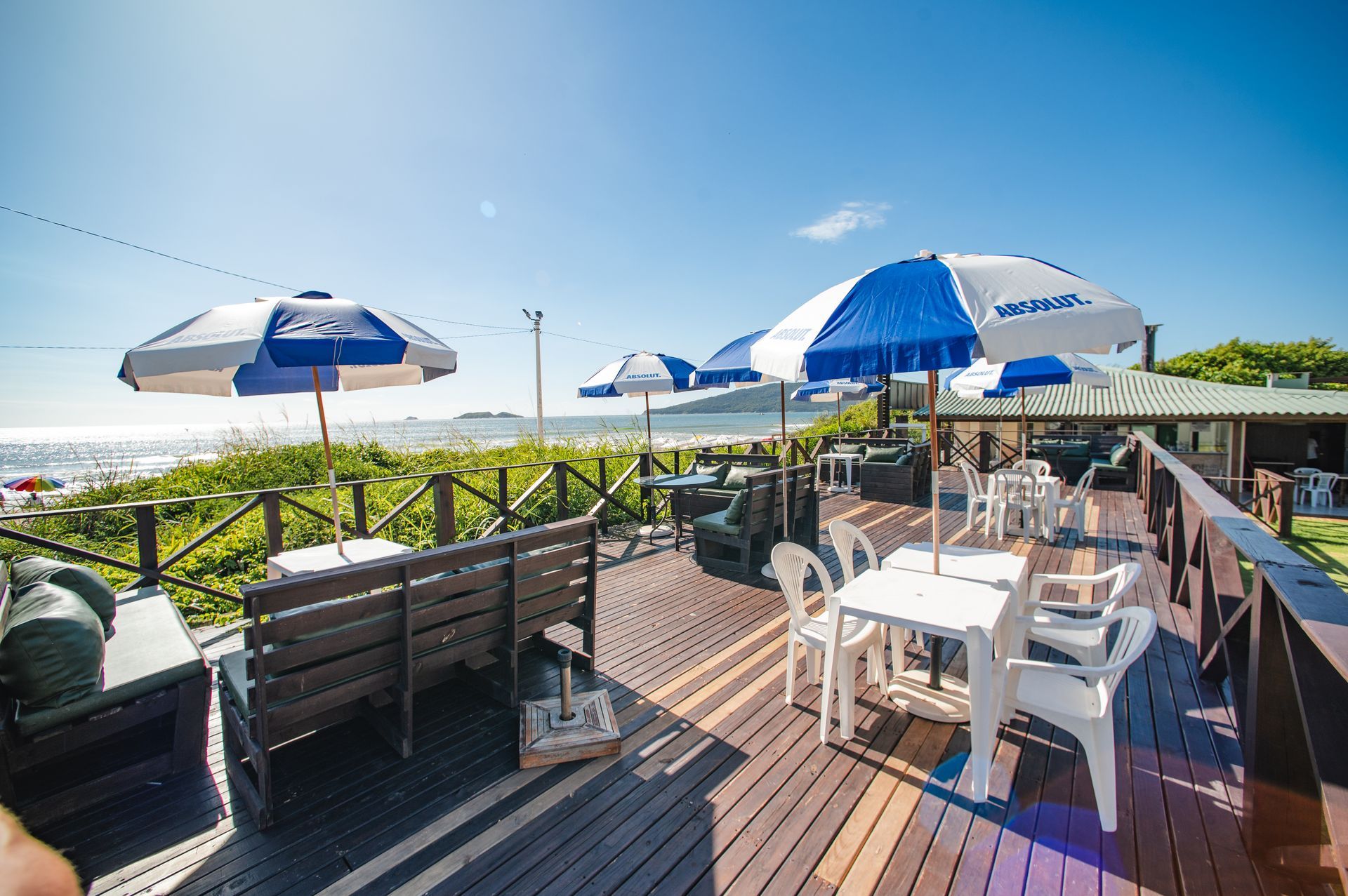 Terraço ensolarado ao ar livre com mesas brancas e guarda-sóis azuis com vista para o oceano.