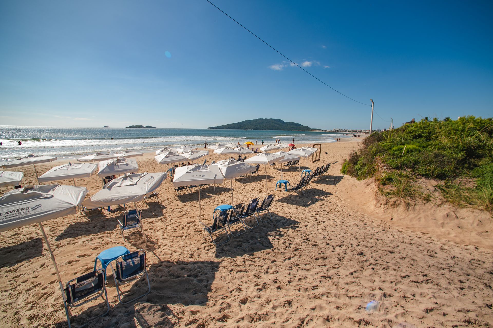 Praia de areia com espreguiçadeiras brancas, guarda-sóis azuis, ondas do oceano e um céu azul límpido.