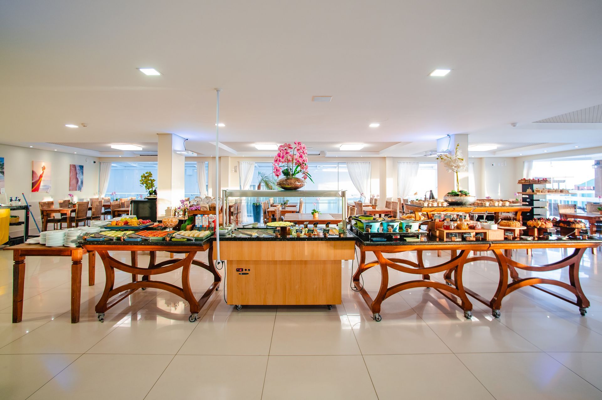 Área de refeições tipo bufê em hotel, bem iluminada, com mesas de madeira, estações de comida e piso de azulejos brancos.